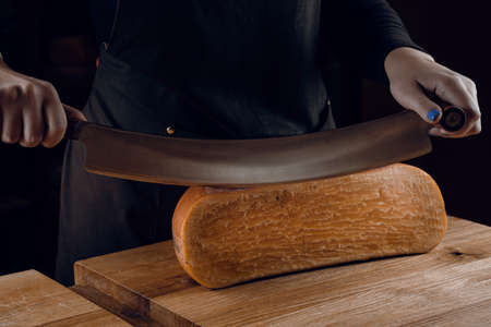 Slicing aged cheese parmesan with crystals using a cheesy dutch knife. Hard cheese with knife on dark background. Snack tasty piece of food for appetizerの写真素材