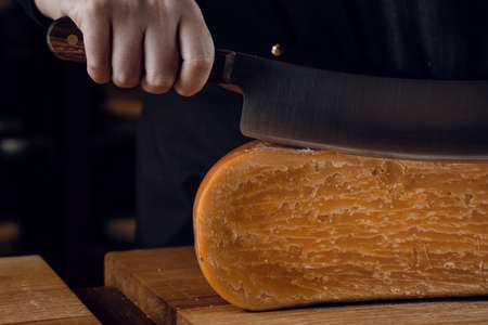 Close-up slicing aged cheese parmesan with crystals using a cheesy dutch knife. Hard cheese with knife on dark background. Snack tasty piece of food for appetizerの写真素材