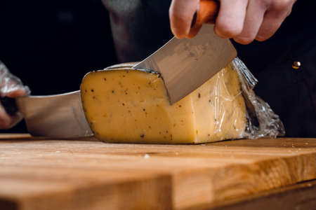 Slicing aged cheese parmesan with crystals using a slicer knife. Hard cheese with knife on dark background. Snack tasty piece of food for appetizerの写真素材