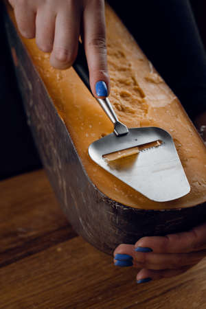 Slicing aged cheese parmesan with crystals using a slicer knife. Hard cheese with knife on dark background. Snack tasty piece of food for appetizerの写真素材