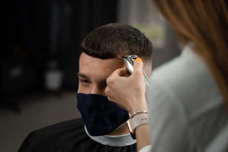 Barbershop service at virus period. Woman barber making hair for handsome man. Using maskの写真素材