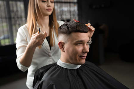 Woman barber making hairstyle in barbershop using clipper. Hairdresser cutting hair of handsome manの写真素材