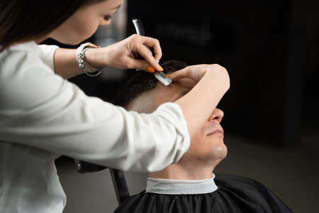 Straight razor cut man hair in barbershop. Attractive woman barber making hairstyle for handsome manの写真素材