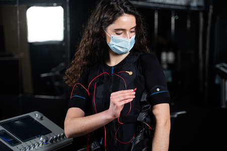 Girl in EMS suit and medical mask in gym. Protection from coronavirus covid-19. Sport training in electrical muscle stimulation suit at quarantine periodの写真素材