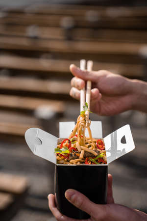 Wok in box in black food container. Holding spicy noodles with chopsticks. Fast food delivery...