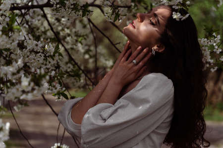The sun shines on the girl's face near the flowering trees in the park. Portrait of attractive girl with curly hair in the gardenの写真素材