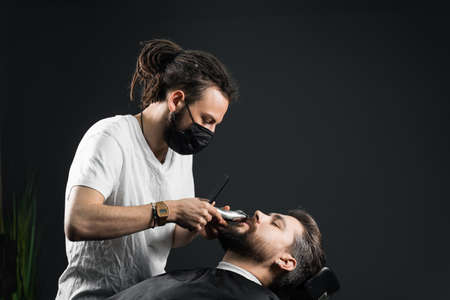 Mustache shaving in barbershop. Barber with dreadlocks in black medical mask trim beard of handsome man at quarantineの写真素材