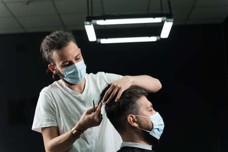 Barber in medical mask is making hairstyle for handsome bearded customer in barbershopの写真素材