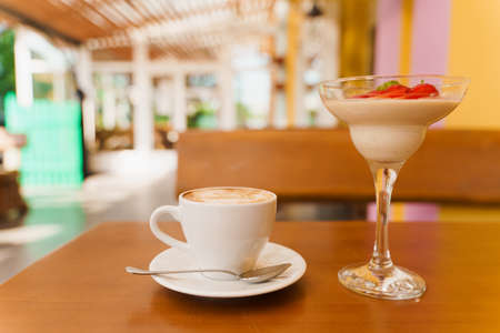 Creamy dessert with strawberries in glass, cappuccino on wooden table. Appetizing dessert and latte on the background of the restaurantの写真素材