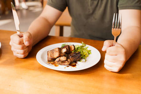 Grill ribs with salad on a white plate on a wooden table. Hungry visitor holding a fork and knifeの写真素材