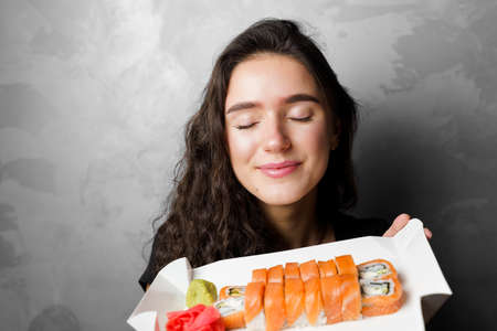 Surprised girl with sushi set philadelphia rolls in a paper box happy girl holding on a gray...
