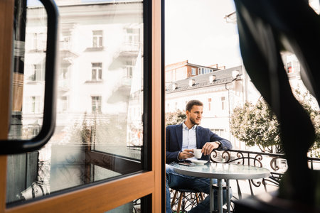 Man is sitting on the summer terrace in cafe, drinking coffee and smiling. Handsome man with cup of coffee in cafe. Morning lifestyle of maleの写真素材