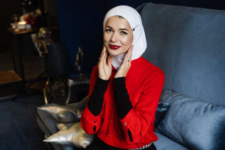 Attractive muslim caucasian girl weared head scarf is sitting on the sofa. Islam religion