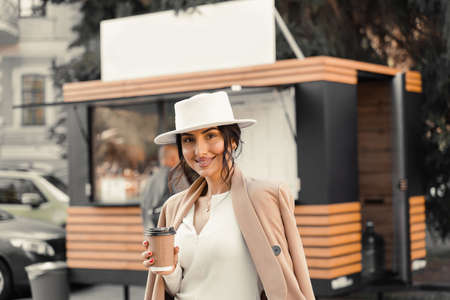 Business lady is drinking coffee near coffee shop. Girl weared white hat, dress and jacketの写真素材