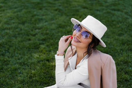 Lifestyle portrait of fashion girl weared white hat, dress, jacket and sunglasses . Woman is sitting on the grassの写真素材