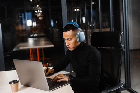 Handsome asian student writes notes and doing distance learning online. Remote work for programmers with laptop and headphonesの写真素材