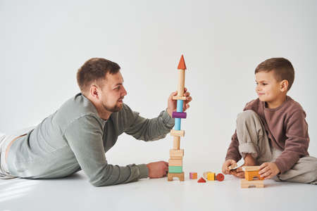 Caring dad helps his son to play on the floor on white background. Father and child build tower of colorful wooden bricks and have fun togetherの写真素材
