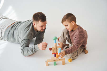 Caring dad helps his son to play on the floor on white background. Father and child build tower of...