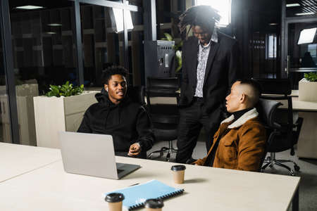 Multiethnic group of African and Asian colleagues are working on laptop on business project. Two black handsome managers teamwork with an Asian man and create creative ideasの写真素材