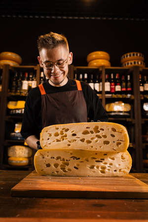 Cheese sommelier in food shop worker holding 2 big pieces of cheese wheel of limited maasdam natural aged. Handsome bearded man working in food storeの写真素材