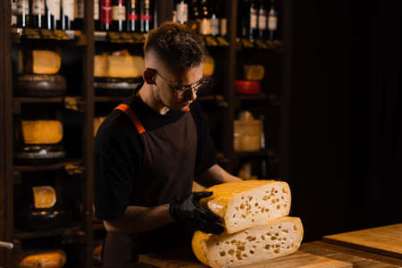 Cheese sommelier in food shop worker holding 2 big pieces of cheese wheel of limited maasdam natural aged. Handsome bearded man working in food storeの写真素材