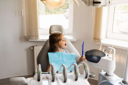 Teeth treatment. Child looking in the mirror at the dentist. Happy child patient of dentistry. Attractive kid girl sitting in dental office and smilingの写真素材