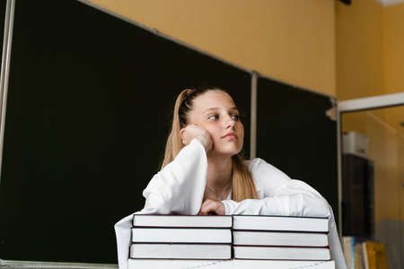 Tired and frustrated schoolgirl lies on books at blackboard in school. School education. The girl is tired of teaching homeworkの写真素材