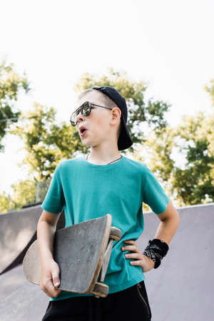 Fashion child boy in glasses posing with skate board on sport playground. Active teenager with sport equipment on sport ramp. Extreme lifestyleの写真素材