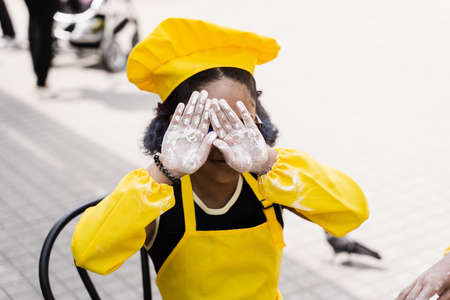 Black african cook child girl showing hands with flour and close her face. African child in chefs hat and yellow apron uniformの写真素材