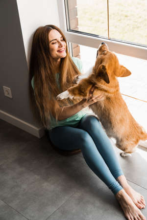 Happy girl hug Corgi dog at home. Lifestyle with domestic pet. Young woman sitting and play with Welsh Corgi Pembrokeの写真素材