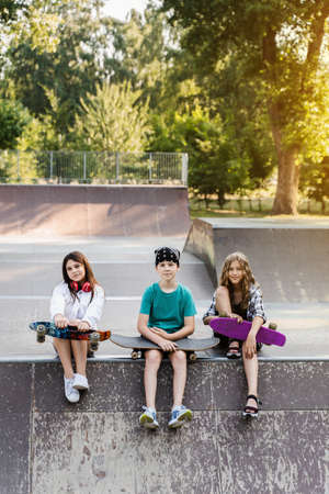 Phone addicted sports kids with skateboard and penny boards are sitting and looking at smartphones on sports ramp on playground. Children addiction of phones.の写真素材