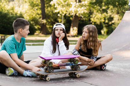 Phone addicted sports kids with skateboard and penny boards are sitting and looking at smartphones on sports ramp on playground. Children addiction of phones.の写真素材