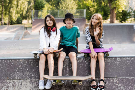 Phone addicted sports kids with skateboard and penny boards are sitting and looking at smartphones on sports ramp on playground. Children addiction of phones.の写真素材