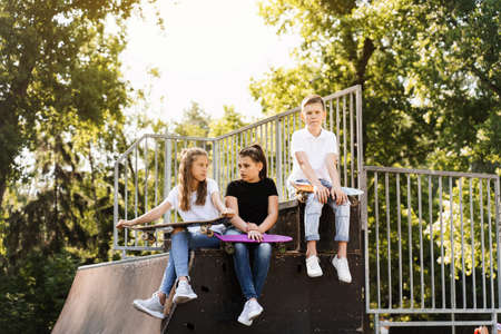 Phone addicted sports kids with skateboard and penny boards are sitting and looking at smartphones on sports ramp on playground. Children addiction of phones.の写真素材