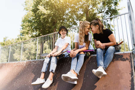 Phone addicted sports kids with skateboard and penny boards are sitting and looking at smartphones on sports ramp on playground. Children addiction of phones.の写真素材