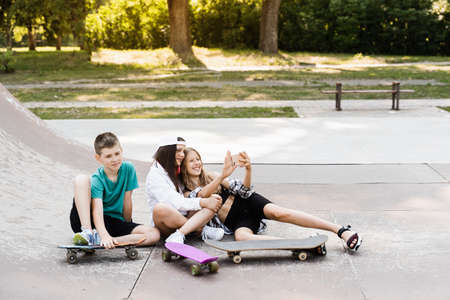 Phone addicted sports kids with skateboard and penny boards are sitting and looking at smartphones on sports ramp on playground. Children addiction of phones.の写真素材