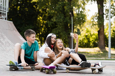 Phone addicted sports kids with skateboard and penny boards are sitting and looking at smartphones on sports ramp on playground. Children addiction of phones.の写真素材