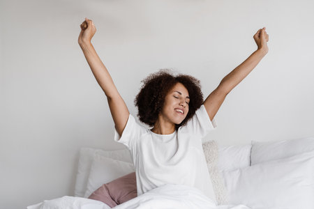 African girl in pajamas stertching in cozy bed and raising hands up at home. African american woman with closed eyes wake up and enjoying good morningの写真素材