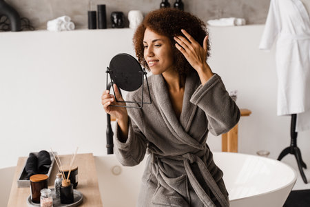 Beauty morning routine of african girl in bathroom. Attractive african american woman in bathrobe is looking in the mirrorの写真素材