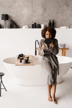 African girl in bathrobe is looking in the mirror on her face in white bathroom. African american cheerful woman doing beauty spa procedures and take skin careの写真素材