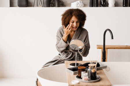 African american woman in bathrobe with facial moisturizing cream doing morning beauty routine. African girl applying face moisturizer cream to protect skin from dryness in the bathの写真素材