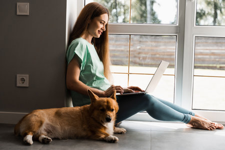 Pregnant girl with laptop and Corgi dog sitting on the floor and chatting online with friends. Lifestyle pregnancy with Welsh Corgi Pembrokeの写真素材