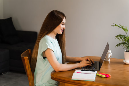 Cheerful girl with laptop typing text and chatting with friends and family in at home. Young woman...