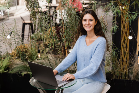 Attractive girl with laptop is sitting in cafe outdoor and chatting with friends and colleagues. Young woman is working on laptop remotely outdoor in restaurantの写真素材