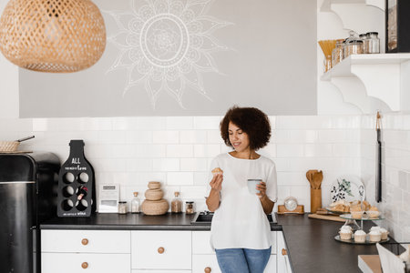 Cozy morning of african american girl on the kitchen at home. Attractive african woman with coffee and cake on the white kitchenの写真素材
