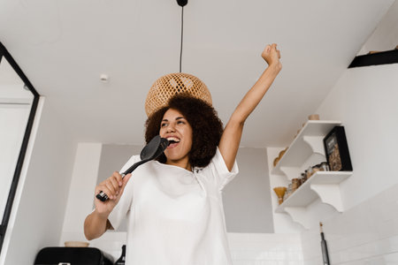 African american housewife in apron singing with spatulas for cooking microphone on the kitchen. Joyful African young woman having fun with kitchen utensilsの写真素材