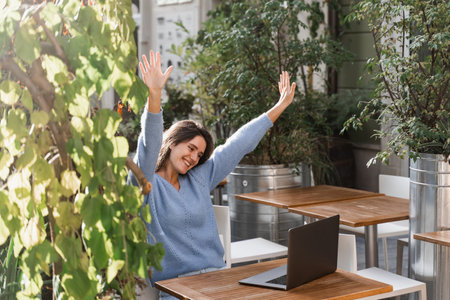 Happy girl with laptop is chatting and greeting with colleagues via video connection outdoor in cafe on terrace. Cheerful young woman is having fun online with friends and familyの写真素材