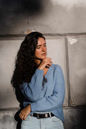 Portrait of attractive business woman with curly hair is posing on grey wall background. Confident girl model weared in blue pullover is posing outdoor in cityの写真素材