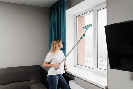 Woman cleaner with mop is cleaning window at home. Windows cleaning service. Housewife is doing...