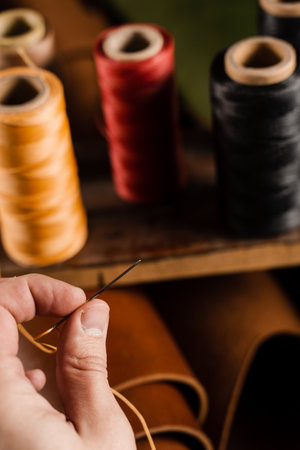 Craftsman is holding needle with thread on roll of brown genuine leather background in craftsman...